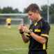 Jeune arbitre sportif en tenue noire et jaune examinant son sifflet sur un terrain de football amateur avant le début d’un match.