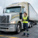Conductrice routière en gilet de sécurité à côté d’un camion poids lourd sur une plateforme logistique du Grand Est.