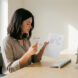 Jeune professionnelle assise dans un bureau moderne, comparant deux documents près de son ordinateur sous une lumière naturelle.