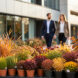 Massif fleuri composé de vivaces en godets devant un bâtiment d’entreprise moderne, avec des employés flous en arrière-plan sous une douce lumière d’octobre.